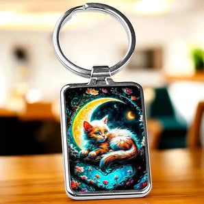 Porte-clés rectangulaire argenté avec un chat couché sur un croissant de lune, décoré de fleurs et d'étoiles.